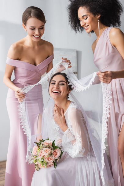 happy multicultural women covering laughing bride with veil in bedroom - 写真・画像
