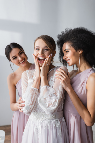 amazed bride touching face while looking at camera near multicultural bridesmaids - 写真・画像
