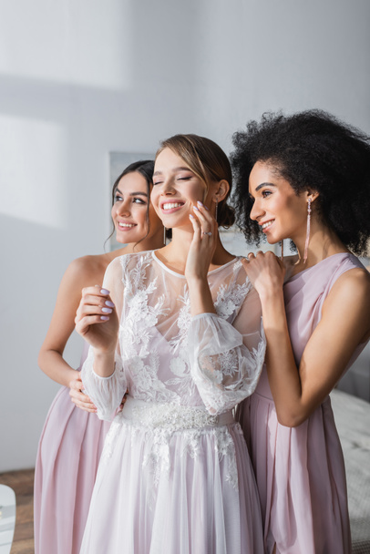 happy bride with closed eyes touching face near smiling interracial bridesmaids - Foto, imagen