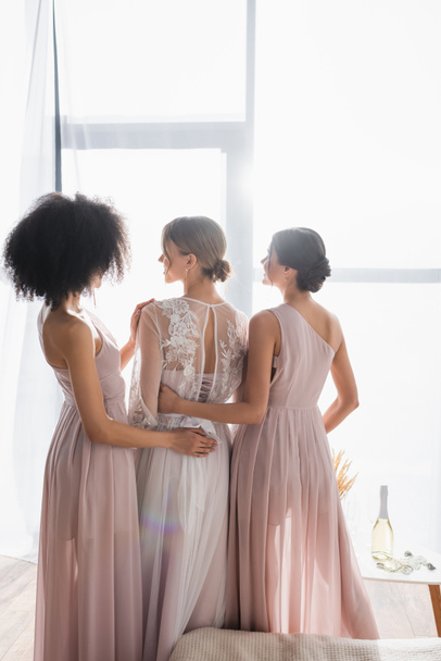 back view of bride near multicultural bridesmaids hugging her in bedroom - Photo, Image