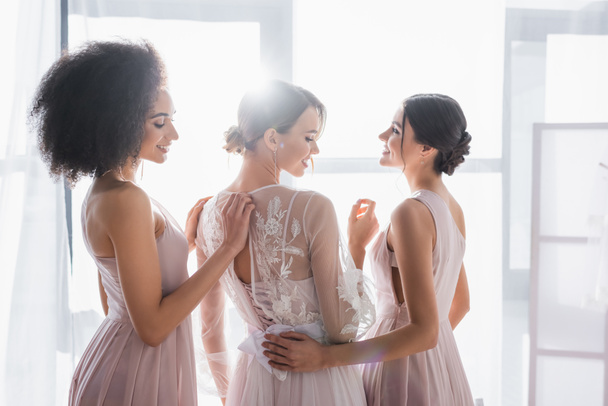 young multiethnic bridesmaids standing near charming bride in sunshine at home - Foto, imagen