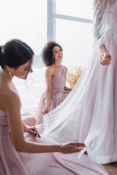 interracial bridesmaids preparing woman for wedding at home, blurred foreground - Foto, afbeelding