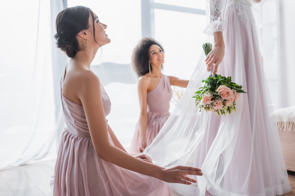 happy multicultural bridesmaids looking at bride holding wedding bouquet - Фото, изображение