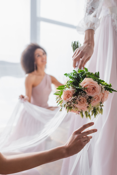 selective focus of wedding bouquet in hand of bride near african american bridesmaid on blurred background - Фото, зображення