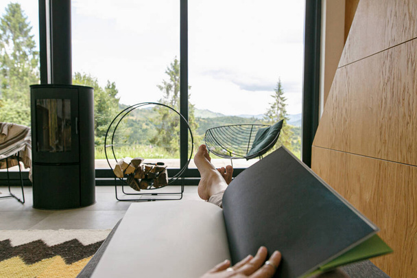 Beautiful female feet and book on background of  fireplace and big window in modern chalet. Young woman barefoot relaxing with book in comfortable home, cozy vacation. Leisure time - Photo, Image