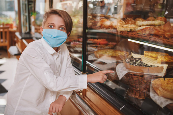 Young boy wearing medical face mask looking shocked while picking dessert to buy at the bakery - 写真・画像