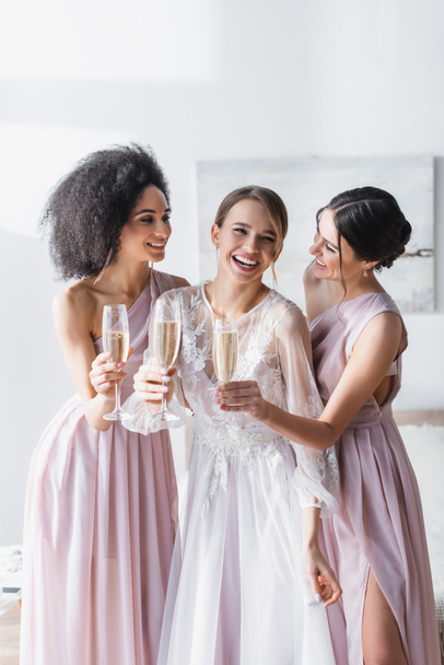 lachende Braut hält Champagner neben fröhlichen interrassischen Freunden im Schlafzimmer - Foto, Bild