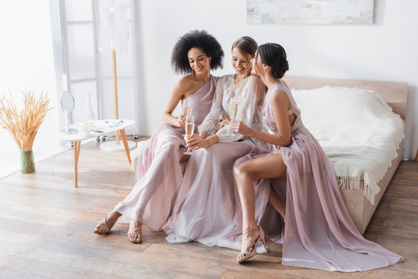 elegant bride with multicultural bridesmaids holding champagne glasses in bedroom - Photo, Image