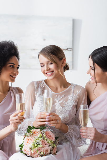 damas de honor multiculturales mirando a la novia feliz mientras sostiene copas de champán - Foto, Imagen