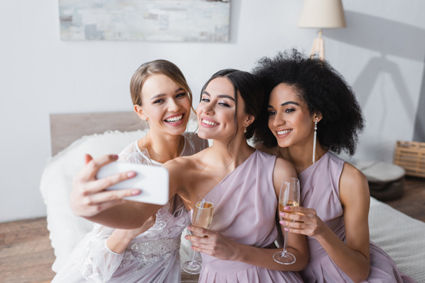 cheerful woman taking selfie with bride and african american friend in bedroom - Фото, изображение