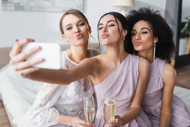 pretty bride with multicultural bridesmaids blowing air kisses while taking selfie on blurred foreground - Photo, Image