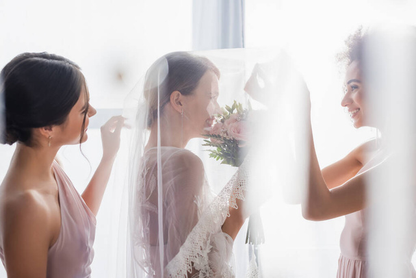 zijaanzicht van gelukkige interraciale bruidsmeisjes in de buurt glimlachende bruid in sluier op wazig voorgrond - Foto, afbeelding