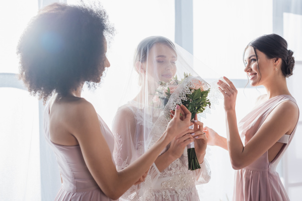 bride holding wedding bouquet with closed eyes while multicultural bridesmaids covering her with veil - Foto, immagini