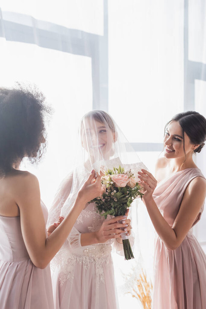 multicultural bridesmaids touching veil of smiling bride holding wedding bouquet - Foto, afbeelding