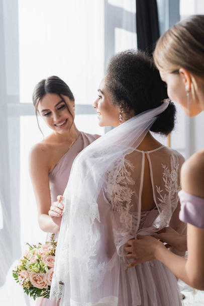 smiling bridesmaids preparing african american bride for wedding, blurred foreground - Photo, Image