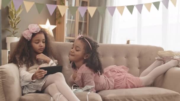 Středně dlouhý záběr kudrnaté Mixed-Race holčička v pěkném oblečení, sedí na gauči v obývacím pokoji, její sestra se převaluje přes pohovku, děti sledují smartphone - Záběry, video