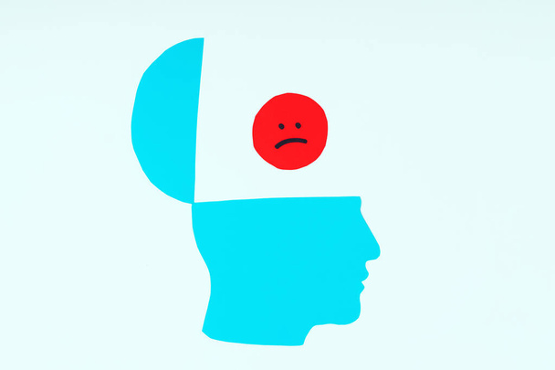 Το χάρτινο προφίλ του μπλε κεφαλιού με μια ελαφρώς ανοιχτή κορυφή με ένα κόκκινο χαρτί λυπημένο χαμογελαστό πρόσωπο. Αρνητικές σκέψεις, ψυχολογία - Φωτογραφία, εικόνα