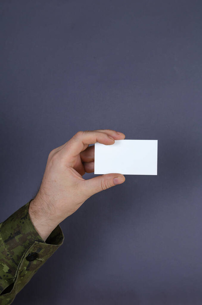 Bir ordu subayının eli gri bir arkaplanda boş bir kartvizit tutuyor. Kamuflaj üniformalı orta yaşlı bir yetişkin. Askeri tema, sözleşmeli askerlik konsepti. - Fotoğraf, Görsel