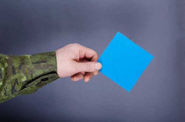 Το χέρι ενός αξιωματικού του στρατού κρατά ένα μπλε τετράγωνο χαρτί σε γκρι φόντο. Ένας μεσήλικας ενήλικας με στολή καμουφλάζ. Στρατιωτικά θέματα, στρατιωτική θητεία, βετεράνοι ιδεών. - Φωτογραφία, εικόνα