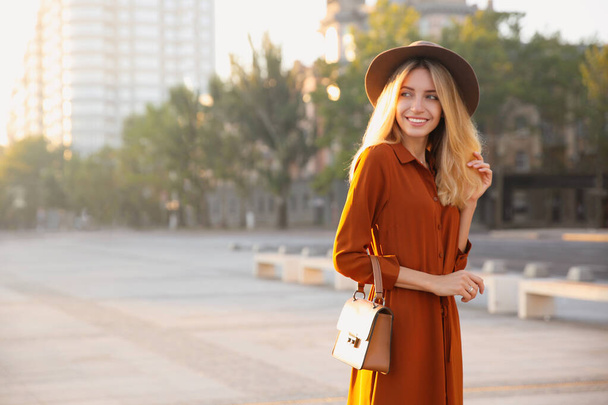 Beautiful young woman in stylish red dress and hat with handbag on city street - Foto, Imagem
