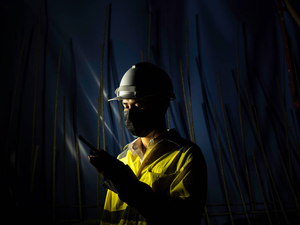 Молодой азиатский инженер в жёлтой куртке безопасности с высокой видимостью, маске для лица и белом защитном шлеме или каске с помощью раций на фоне строительства тёмного железнодорожного тоннеля. - Фото, изображение