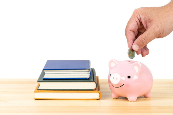 Osoba wkładająca pieniądze do różowego skarbonki na drewnianym biurku z książkami stos na białym tle, oszczędność koncepcji na edukację i stypendium - Zdjęcie, obraz