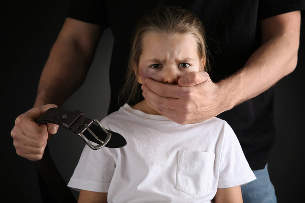 Man with belt covering scared little girl's mouth on black background. Domestic violence - Foto, Imagen