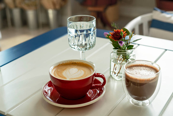 cappuccino in red mug, cacao in glass cup, glass of water and small bouquet flowers in glass jar on table in cafe, decor, hot drinks, still life, coffee time - Photo, Image