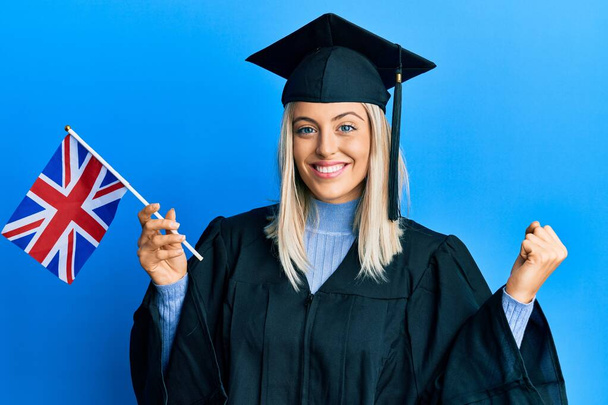 Όμορφη ξανθιά γυναίκα φορώντας καπέλο αποφοίτησης και τελετή ρόμπα κρατώντας σημαία Ηνωμένο Βασίλειο ουρλιάζοντας υπερήφανοι, γιορτάζει τη νίκη και την επιτυχία πολύ ενθουσιασμένος με έθεσε το χέρι  - Φωτογραφία, εικόνα