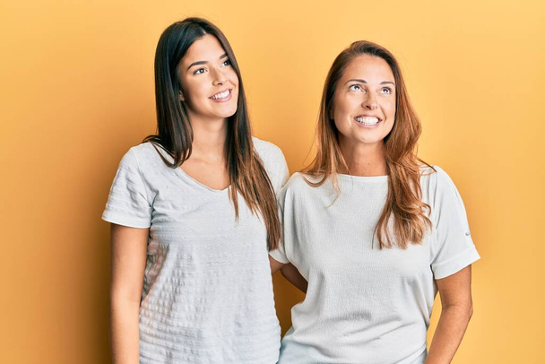 Ισπανόφωνη οικογένεια, μητέρα και κόρη, φορώντας ένα απλό λευκό μπλουζάκι, χαμογελώντας κοιτώντας στο πλάι και κοιτάζοντας αλλού, σκεπτόμενοι.  - Φωτογραφία, εικόνα