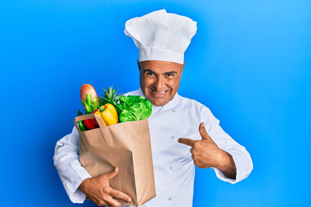 Starší středovýchodní muž profesionální kuchař drží tašku s potravinami ukazující prstem na jedno já usměvavý šťastný a hrdý  - Fotografie, Obrázek