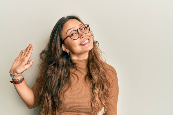 Menina hispânica jovem vestindo roupas casuais e óculos dispensa dizendo olá feliz e sorridente, gesto de boas-vindas amigável  - Foto, Imagem