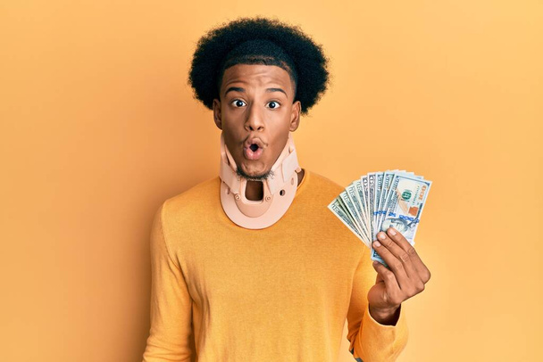 African american man with afro hair wearing cervical neck collar and holding money from insurance scared and amazed with open mouth for surprise, disbelief face  - Фото, изображение
