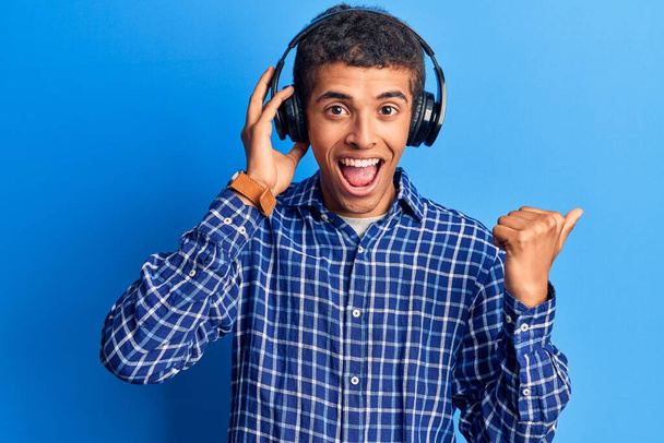 Young african amercian man listening to music using headphones pointing thumb up to the side smiling happy with open mouth  - 写真・画像