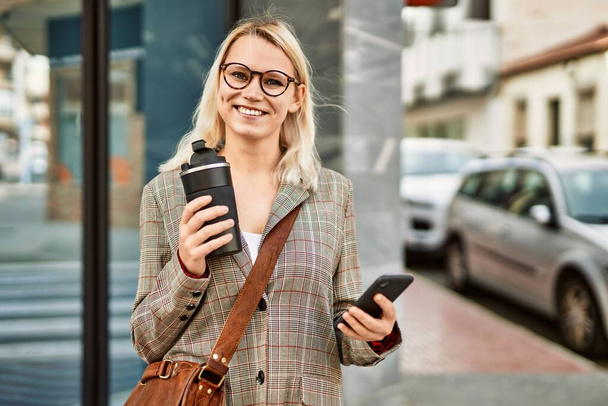 Młoda blondynka bizneswoman za pomocą smartfona pije kawę w mieście. - Zdjęcie, obraz