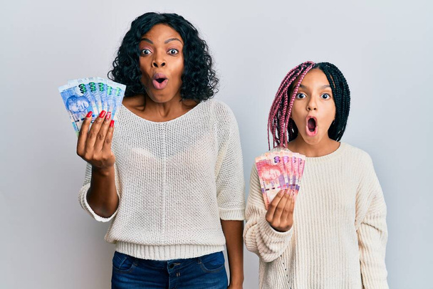 Hermosa madre afroamericana y su hija sosteniendo billetes de rand sudafricanos asustados y sorprendidos con la boca abierta para sorpresa, cara de incredulidad  - Foto, Imagen