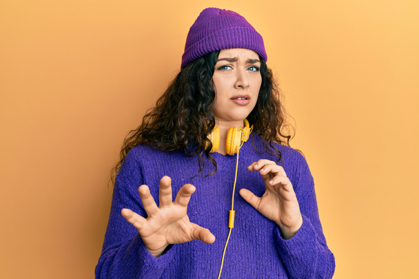 Young brunette woman with curly hair listening to music using headphones disgusted expression, displeased and fearful doing disgust face because aversion reaction. with hands raised  - Foto, Imagen