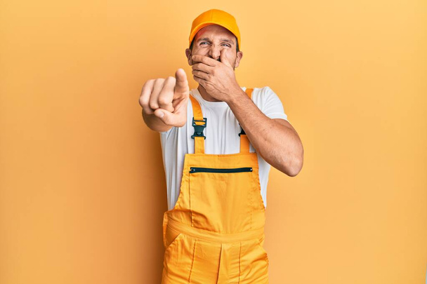 Young handsome man wearing handyman uniform over yellow background laughing at you, pointing finger to the camera with hand over mouth, shame expression  - Photo, Image