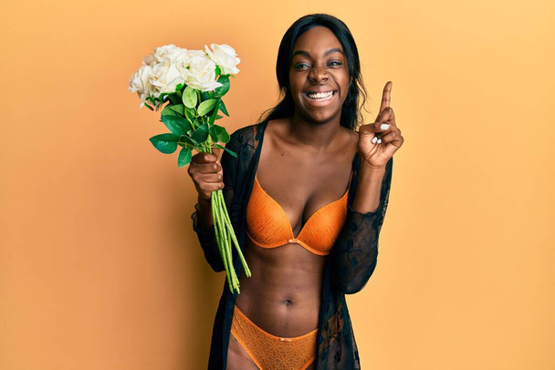 Молода афроамериканська жінка у білизні, тримаючи квіти здивовані ідеєю або питанням, що вказує пальцем на щасливе обличчя, по-перше.  - Фото, зображення