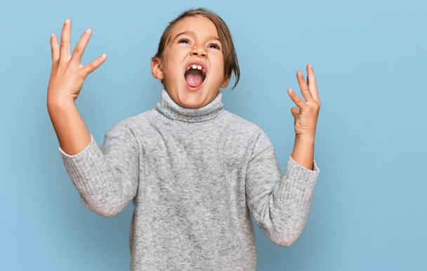 Little beautiful girl wearing casual turtleneck sweater crazy and mad shouting and yelling with aggressive expression and arms raised. frustration concept.  - Фото, зображення