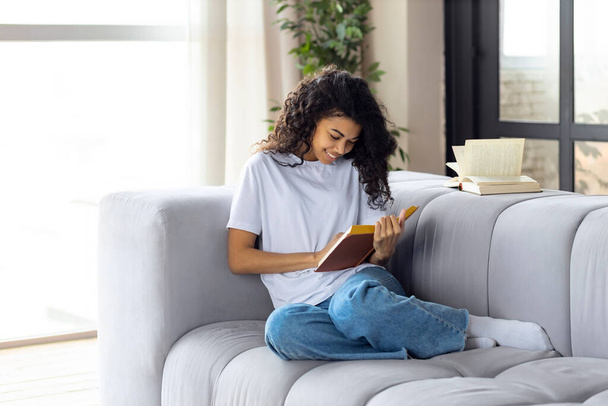 Happy young african american woman reading her favorite book while sitting on sofa at home - Foto, imagen