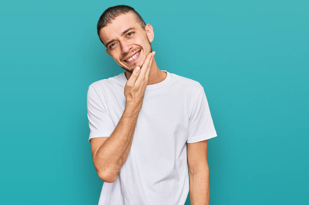 Латиноамериканец в обычной белой футболке уверенно смотрит на камеру, улыбаясь скрещенными руками и поднятой на подбородок рукой. Позитивное мышление.  - Фото, изображение