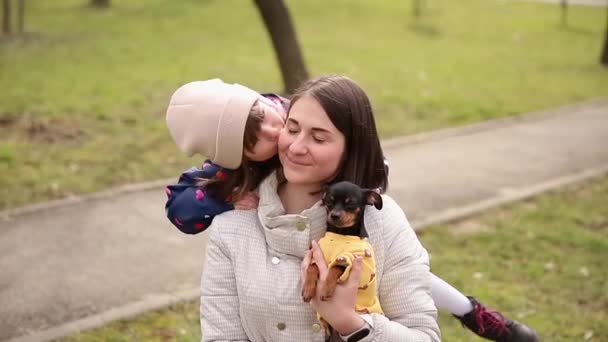 Όμορφη νεαρή μαμά βόλτα με την κόρη και το μικρό σκυλί. Ευτυχισμένη οικογένεια περπατά έξω με το κατοικίδιο ζώο κατά τη διάρκεια της απομόνωσης. Κόρη φιλί μαμά - Πλάνα, βίντεο