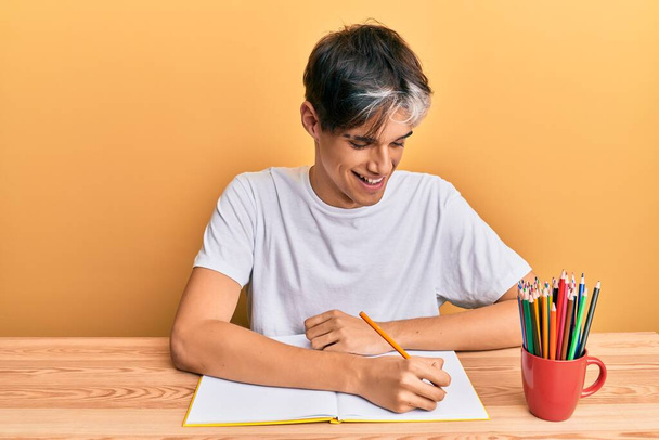 Young hispanic man writing book sitting on the table smiling with a happy and cool smile on face. showing teeth.  - Foto, Imagen