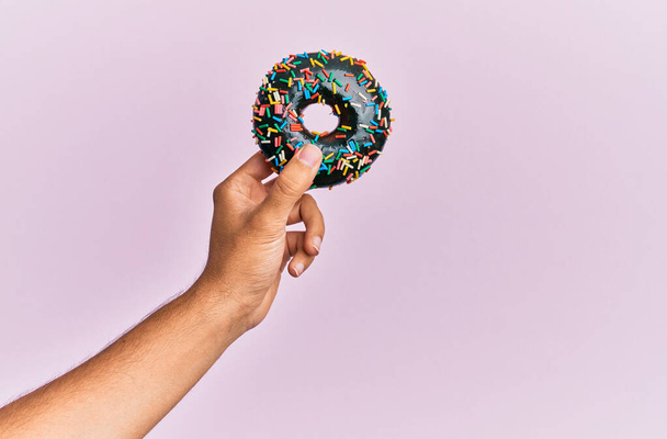 Mão de homem hispânico segurando donut de chocolate sobre fundo rosa isolado. - Foto, Imagem