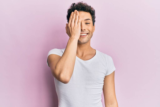 Young handsome man wearing casual white t shirt covering one eye with hand, confident smile on face and surprise emotion.  - Photo, Image