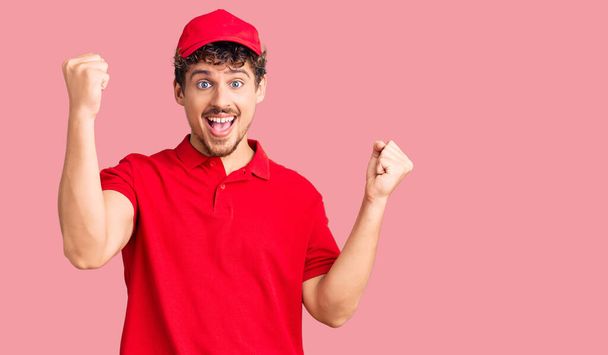 Young handsome man with curly hair wearing delivery uniform screaming proud, celebrating victory and success very excited with raised arms  - Photo, Image