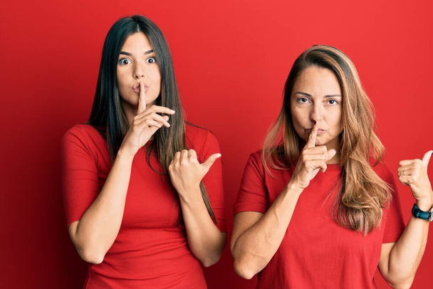 Die hispanische Familie aus Mutter und Tochter trägt lässige Kleidung vor rotem Hintergrund und bittet um Ruhe, wobei der Finger auf den Lippen mit der Hand zur Seite zeigt. Schweigen und geheimes Konzept.  - Foto, Bild