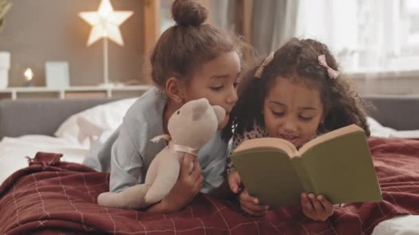 Full shot of two cute Mixed-Race little sisters lying on stomachs in bed, reading book together, smiling - Imágenes, Vídeo