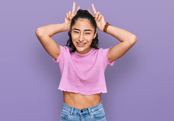 Hispanic teenager girl with dental braces wearing casual clothes posing funny and crazy with fingers on head as bunny ears, smiling cheerful  - Photo, image
